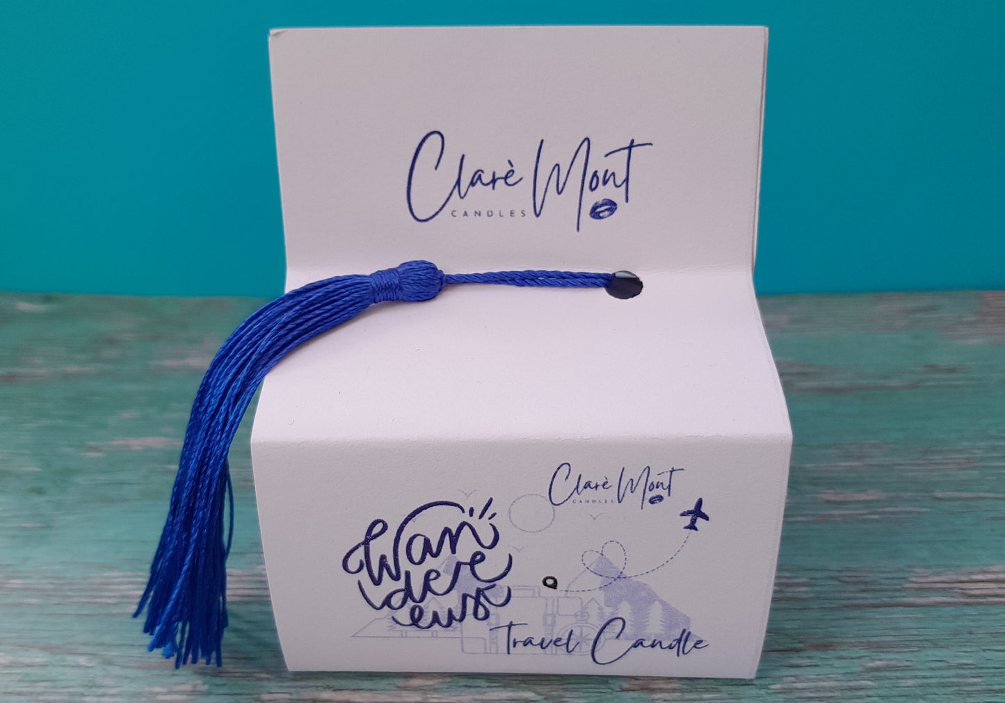 A travel candle with a blue tassel around a white box with 'Clare Mont' branding on a teal background