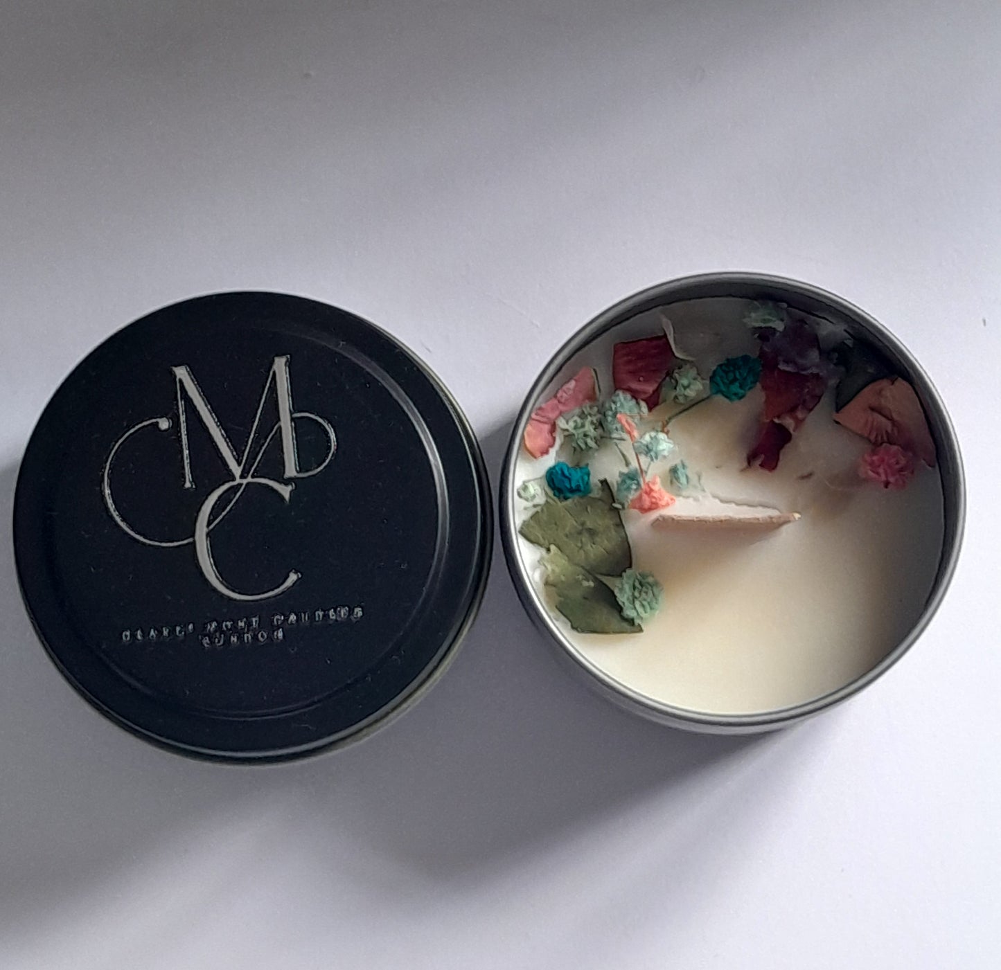 Candle in a tin with decorative petals and a black lid featuring a logo.