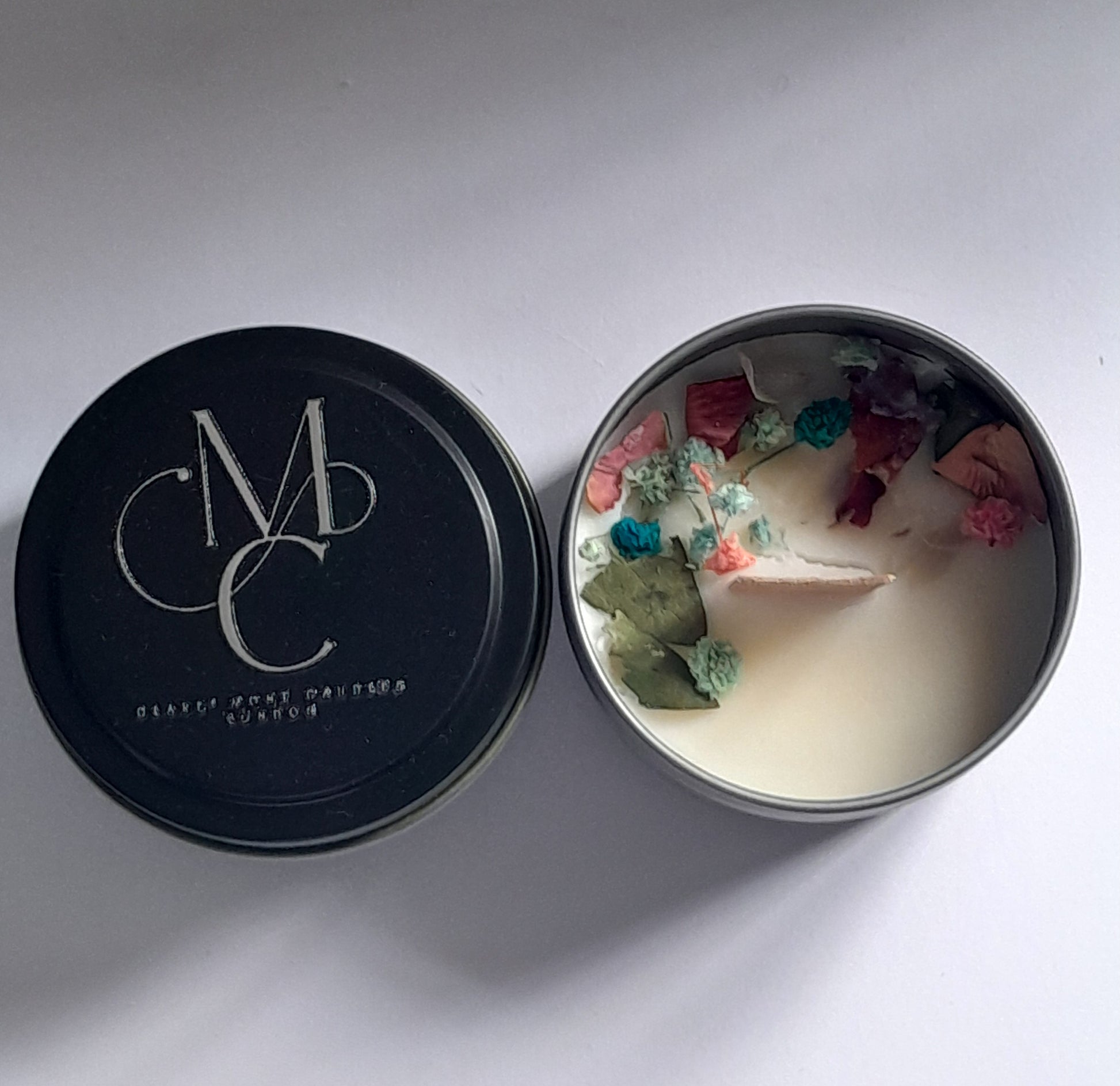 Candle in a tin with decorative petals and a black lid featuring a logo.